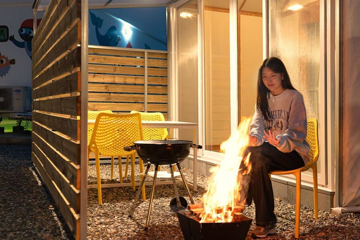 Eine Person sitzt an einem Lagerfeuer in einer Freiluftanlage, mit einem Schild mit der Aufschrift '119 REFRESH GLAMPING 2023'.