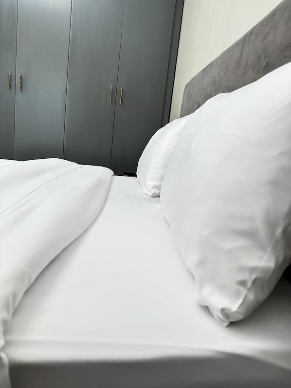 A neatly made bed with white linens and pillows, grey wardrobe with handles, and a grey headboard.