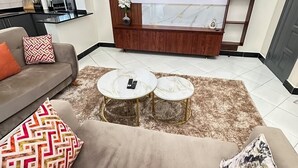 A modern living room with a sofa, two coffee tables, a TV, and a kitchen in the background. - LivLuxe Apartments 2 (Kampala)