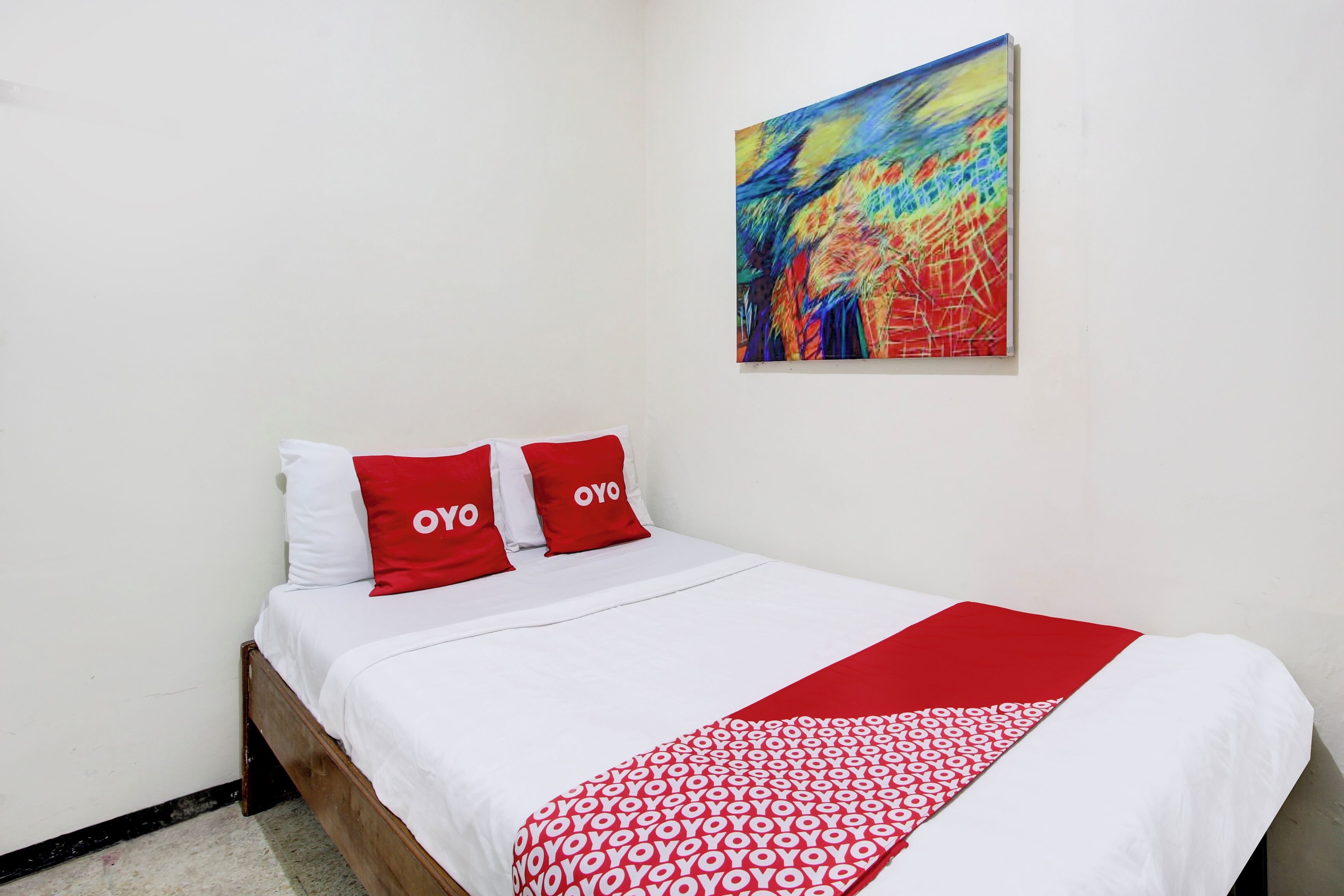 A hotel room with a bed, two red pillows, a white bedspread, a red blanket, and a colorful abstract painting on the wall.