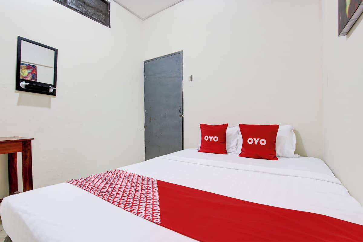 Une chambre d’hôtel avec un lit, deux oreillers rouges ornés du mot « OYO », une couvre-lit blanche et rouge, une table de chevet en bois, un miroir et une porte.
