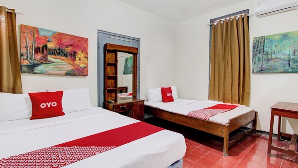 Une chambre d’hôtel avec deux lits, chacun agrémenté d’un oreiller rouge à l’effigie de la marque OYO, d’une table de chevet en bois, d’un miroir et d’un tableau accroché au mur.