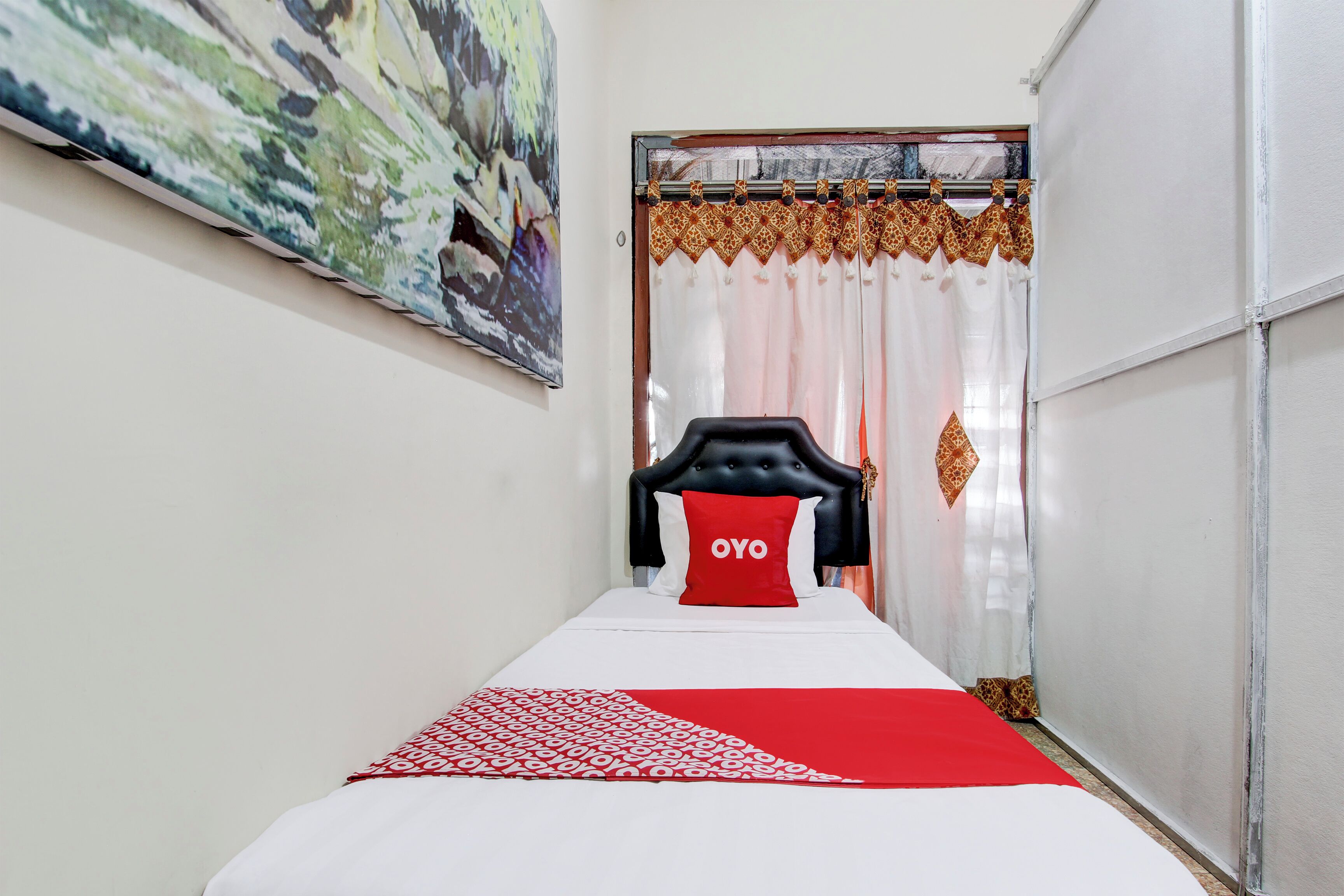 A narrow hotel room with a bed, a headboard, a red pillow, and a patterned bedspread.