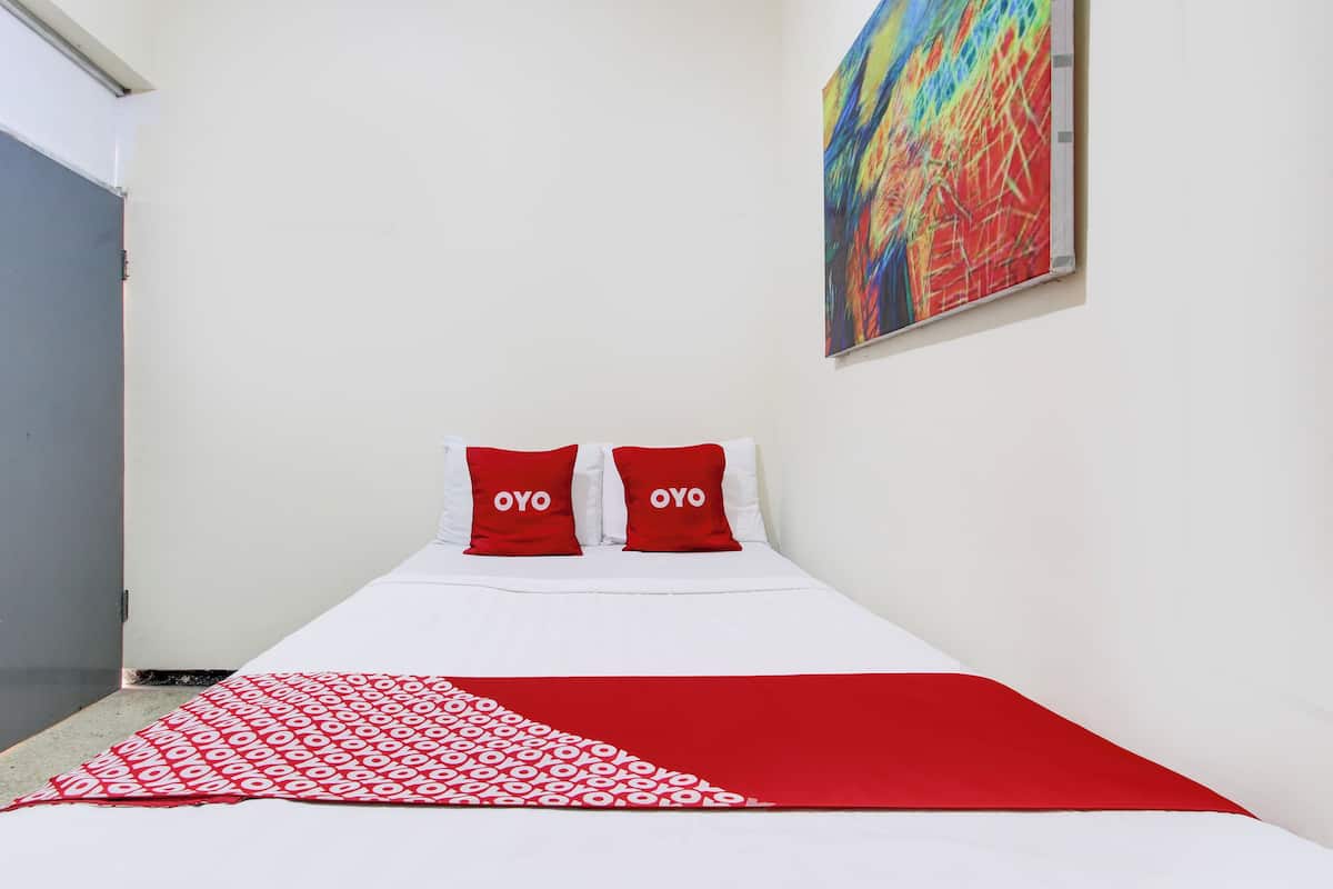 Un lit avec une literie blanche et rouge, deux oreillers rouges ornés du mot « OYO », et un tableau abstrait aux couleurs vives accroché au mur.