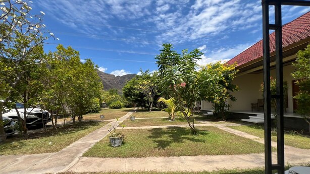 A courtyard with a paved walkway, surrounded by trees and a building with a red roof. - Hotel O Gunung Mas Homestay (Pemuteran)