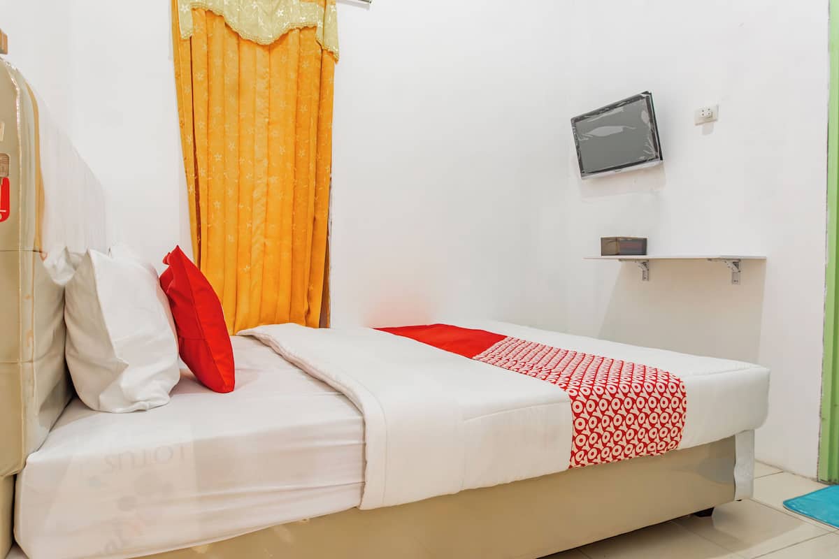 A single bed with a white and red patterned bedspread, a red pillow, and a white pillow. There is a small shelf with a black object on it, and a wall-mounted TV.