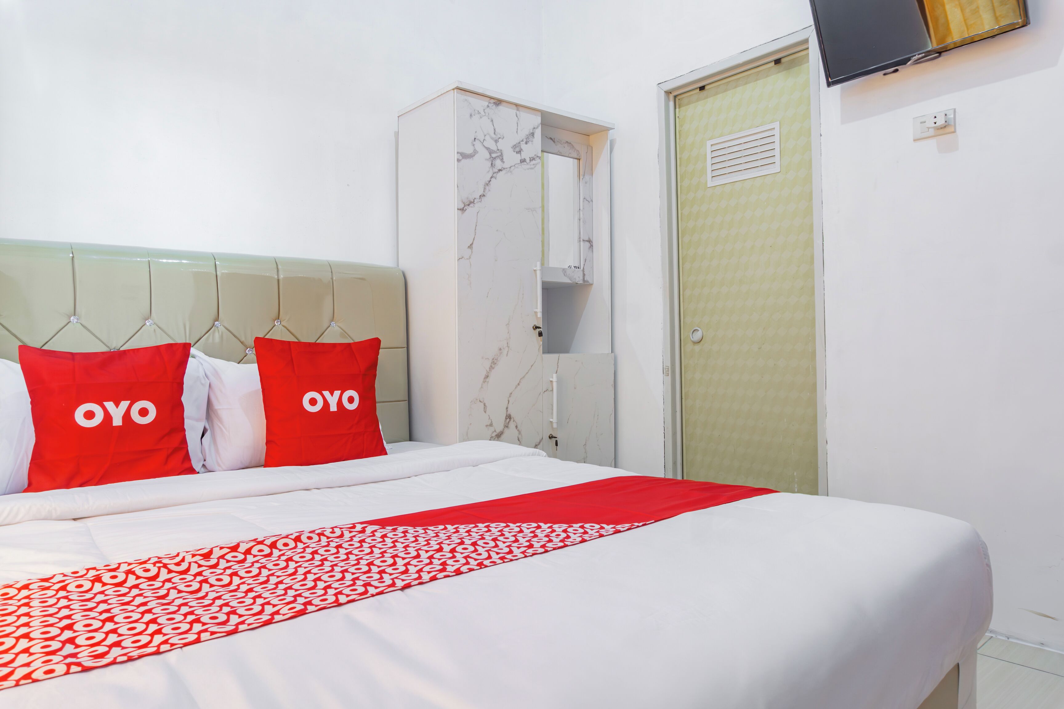 Ein Hotelzimmer mit einem Bett, roten Kissen, einer rot-weißen Bettdecke, einem Marmor-Kopfteil, einem an der Wand befestigten Fernseher und einer Tür mit einem gemusterten Vorhang.