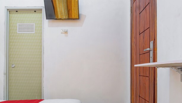 Ein kleines Zimmer mit einem Bett, einem Holzschrank, einer Tür mit Lüftungsgitter und einer Wandleuchte.
