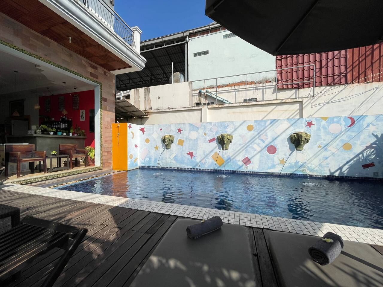 Un espace piscine doté d’une terrasse en bois, de chaises longues et d’une fresque murale aux couleurs vives.