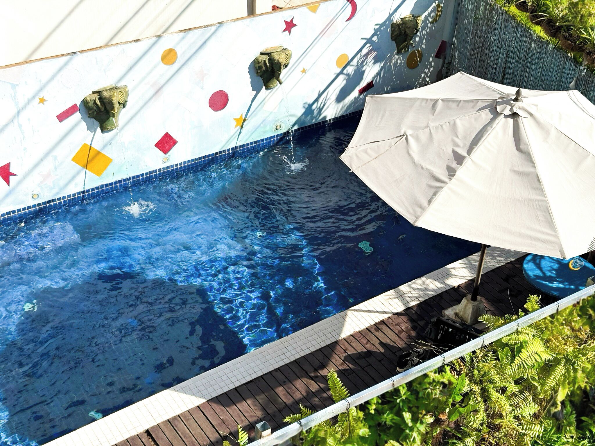 A swimming pool with a tiled edge, a white umbrella, and a wooden deck.