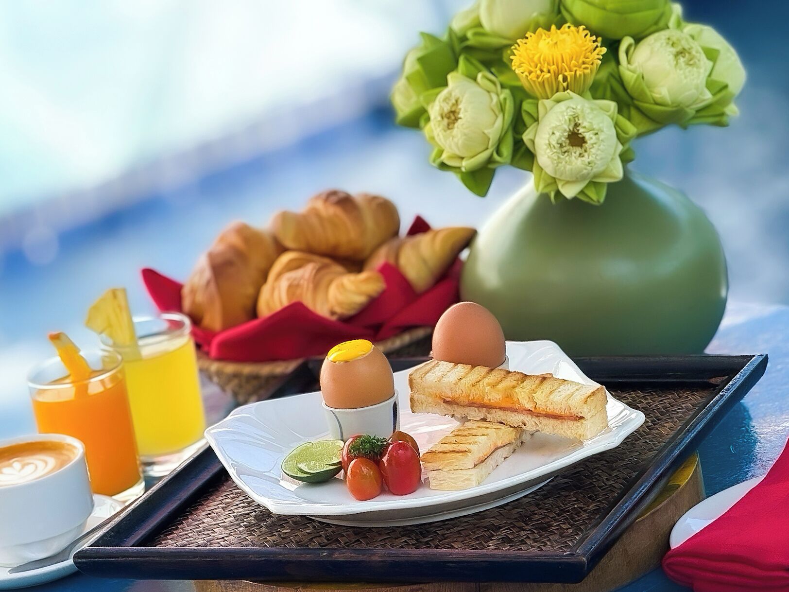 A tray with croissants, a grilled sandwich, a boiled egg, and a drink, accompanied by a vase of flowers and a basket of pastries.