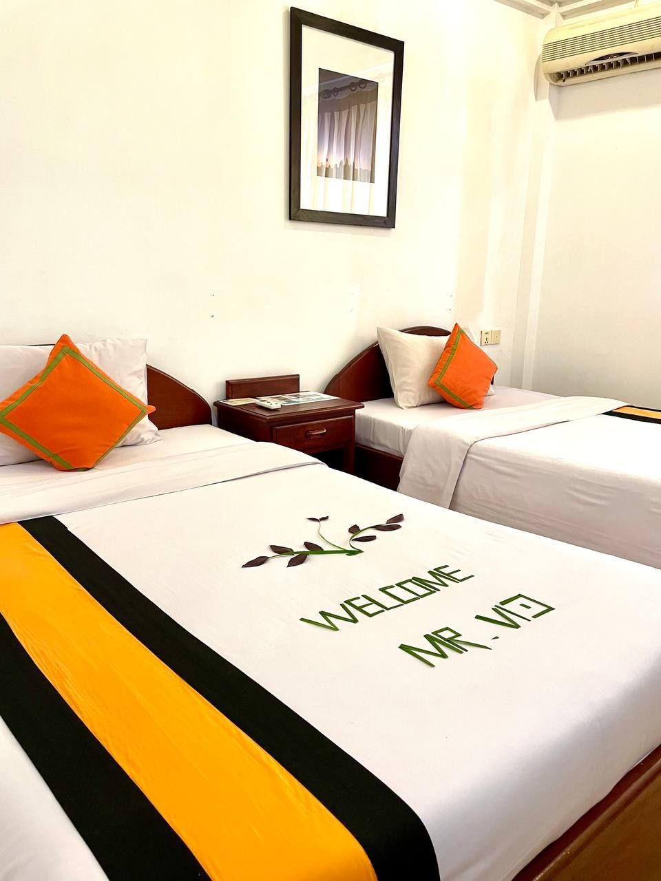 A hotel room with two beds, each with a white bedspread and a 'WELCOME' sign. There is a framed picture on the wall and an air conditioning unit above the beds.