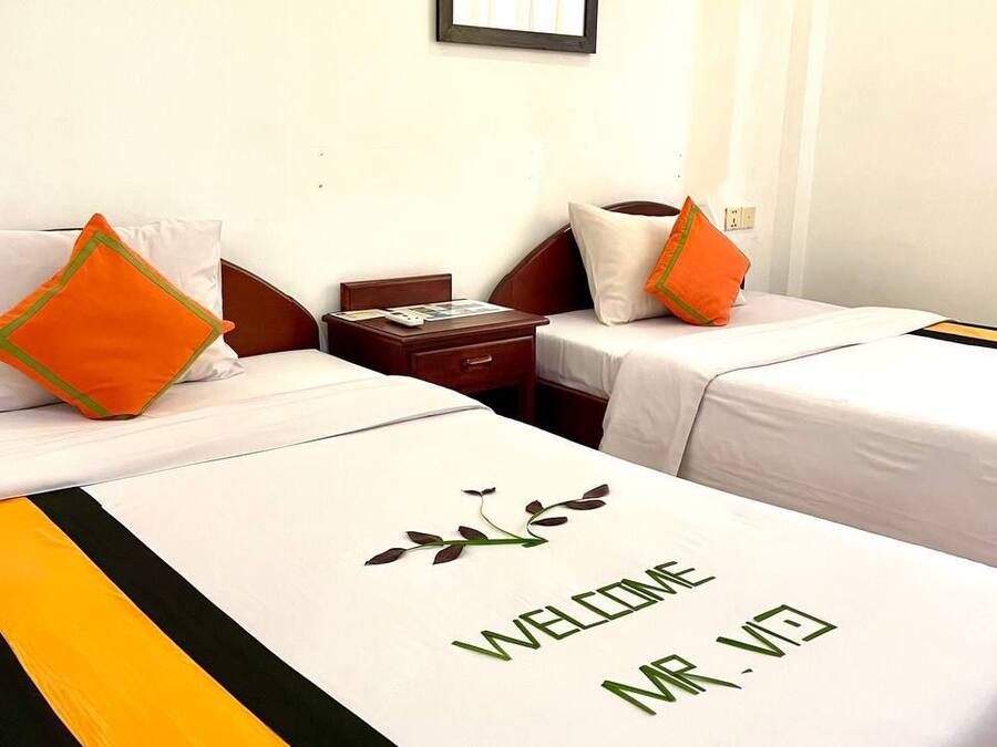 A hotel room with two beds, each with a white bedspread and a 'WELCOME' sign. There is a framed picture on the wall and an air conditioning unit above the beds.