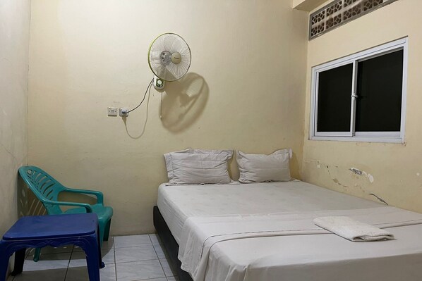 A small, simple room with a bed, a wall-mounted fan, a window, and two plastic chairs. - Capital O 94240 Asoka Syariah (Padang)