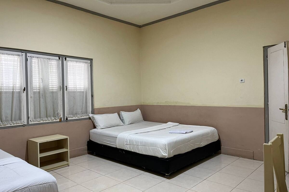 A bedroom with a single bed, a small wooden shelf, and a window with curtains.