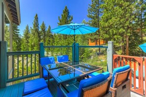 A deck with a glass-top table and blue cushioned chairs, surrounded by a wooden railing and overlooking a forested area.