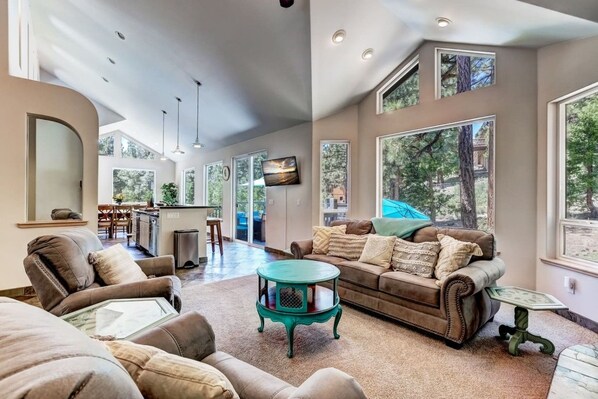 A spacious living room with a large sofa, a coffee table, and a television. There is a kitchen area in the background with a dining table and chairs.