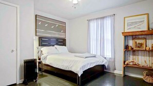 Un dormitorio con cama, un radiador, un ventilador de techo, una ventana con cortinas y una estantería con objetos decorativos.