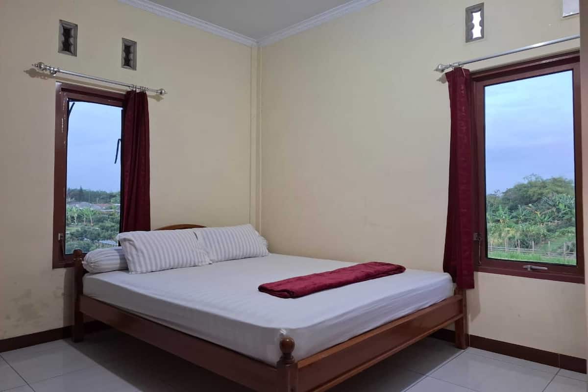 Une chambre avec un lit simple, un cadre en bois, un matelas blanc et une couverture rouge. Il y a deux fenêtres avec des rideaux, une de chaque côté du lit, et la vue donne sur un paysage verdoyant.