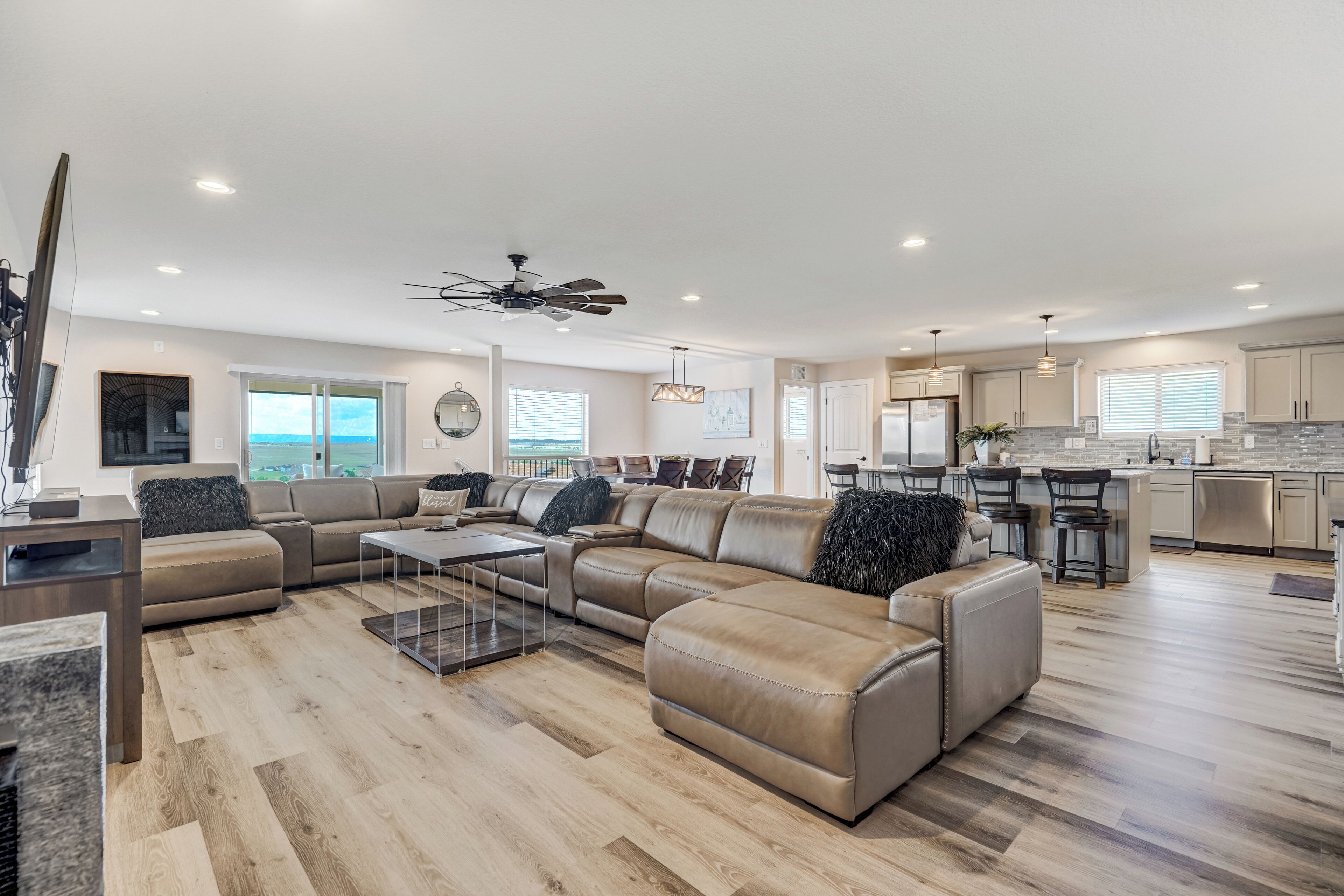 A spacious living room with a large L-shaped sectional sofa, a coffee table, and a dining area with a bar. The kitchen is visible in the background with modern appliances and a large window.