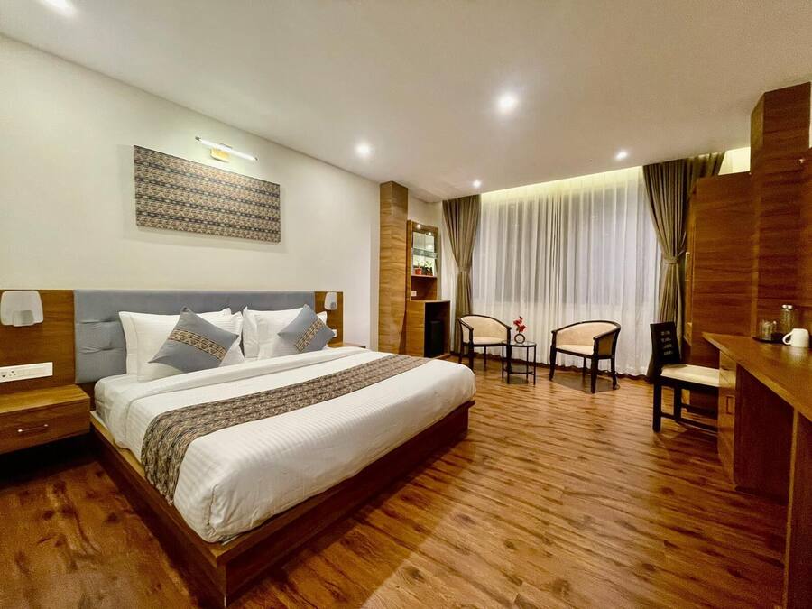 Habitación de hotel con una cama grande, un escritorio con dos sillas, una mesita con un florero y un estante en la pared.
