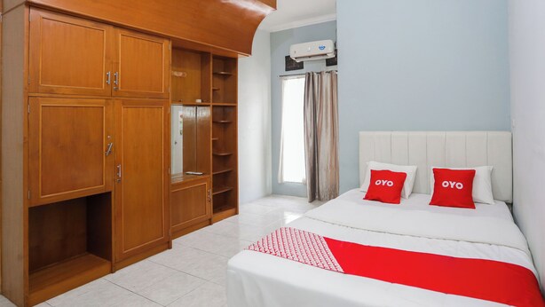 A hotel room with a large wardrobe, a bed with white bedding and red accents, and a window with curtains. - Hotel O Sman House (Bekasi)