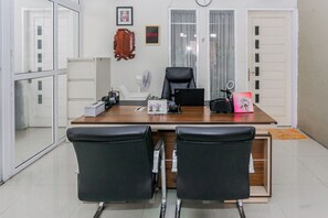 Egy modern iroda, melyben egy fa íróasztal, két fekete bőr szék, egy fehér ventilátor és egy keretezett kép található a falon.