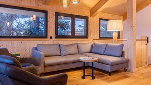 A cozy room with a grey sofa, a small round table, and a lamp. There are large windows with a view of trees and a wooden ceiling.