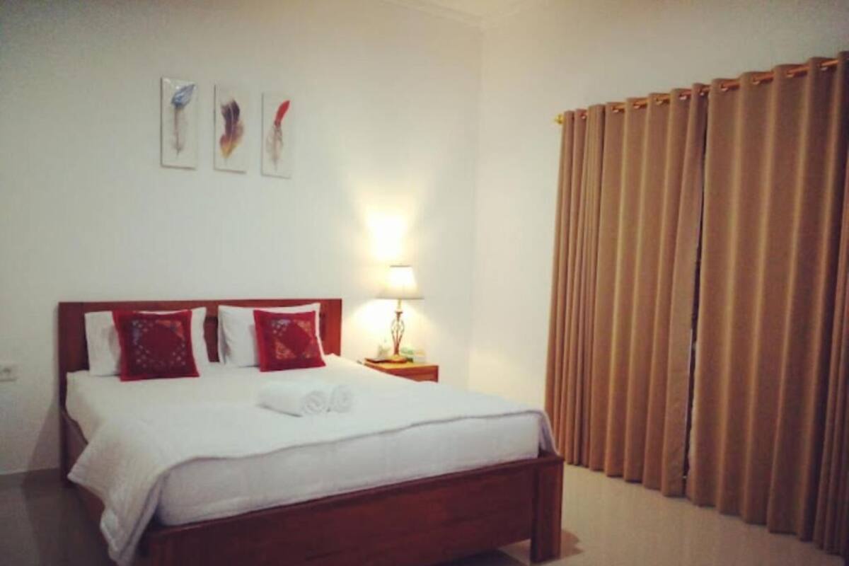 A neatly made bed with white bedding and red pillows, a bedside lamp, and a curtain on the window.