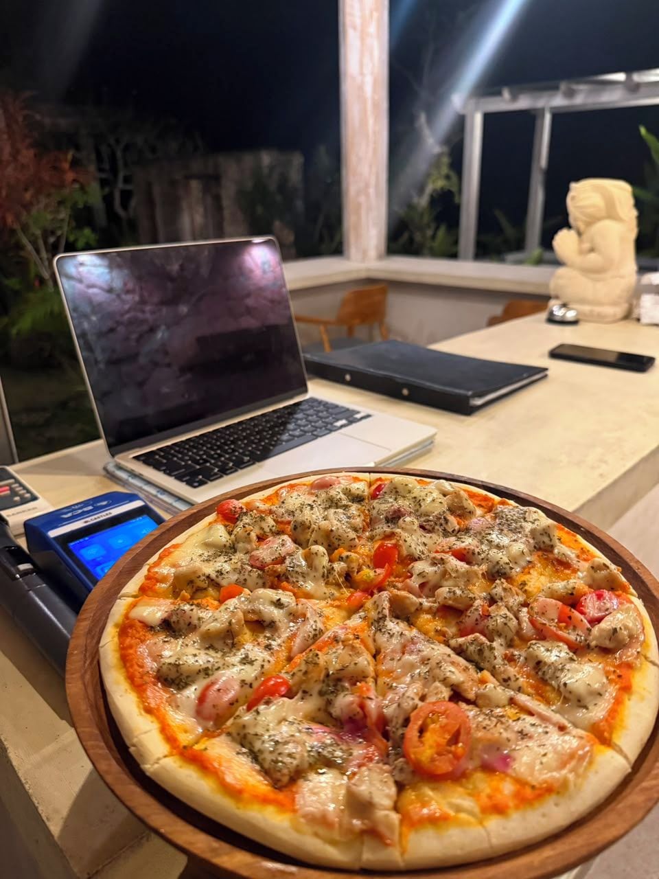 A pizza on a wooden board with a laptop and a smartphone on a table.