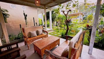 An outdoor seating area with wooden furniture, surrounded by lush greenery and a view of a building in the background.