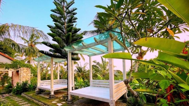 Un jardín con dos camas de madera blanca para colgar bajo una cubierta de vidrio, rodeado de exuberante follaje tropical.