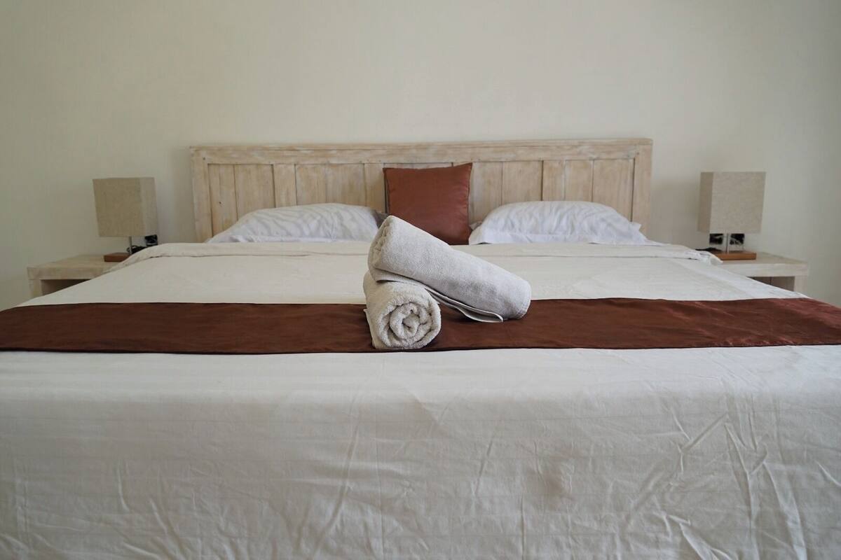 A neatly made bed with white linens, a brown throw blanket, and a rolled towel on the bed.