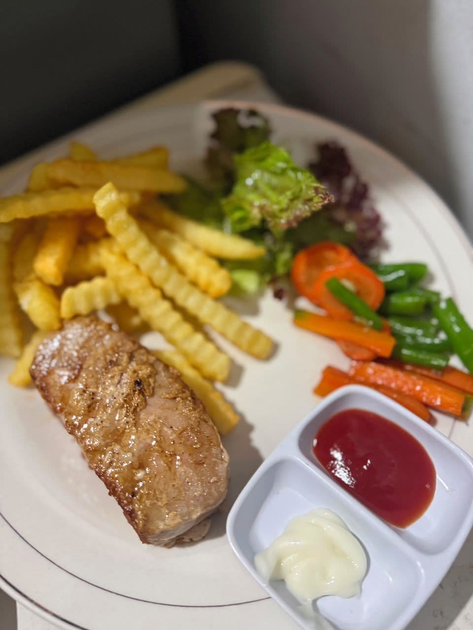 A plate with a piece of meat, French fries, mixed vegetables, and two dipping sauces.