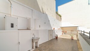 Un estrecho pasillo exterior con un lavamanos blanco, armarios, plantas en macetas y suelo de baldosas.