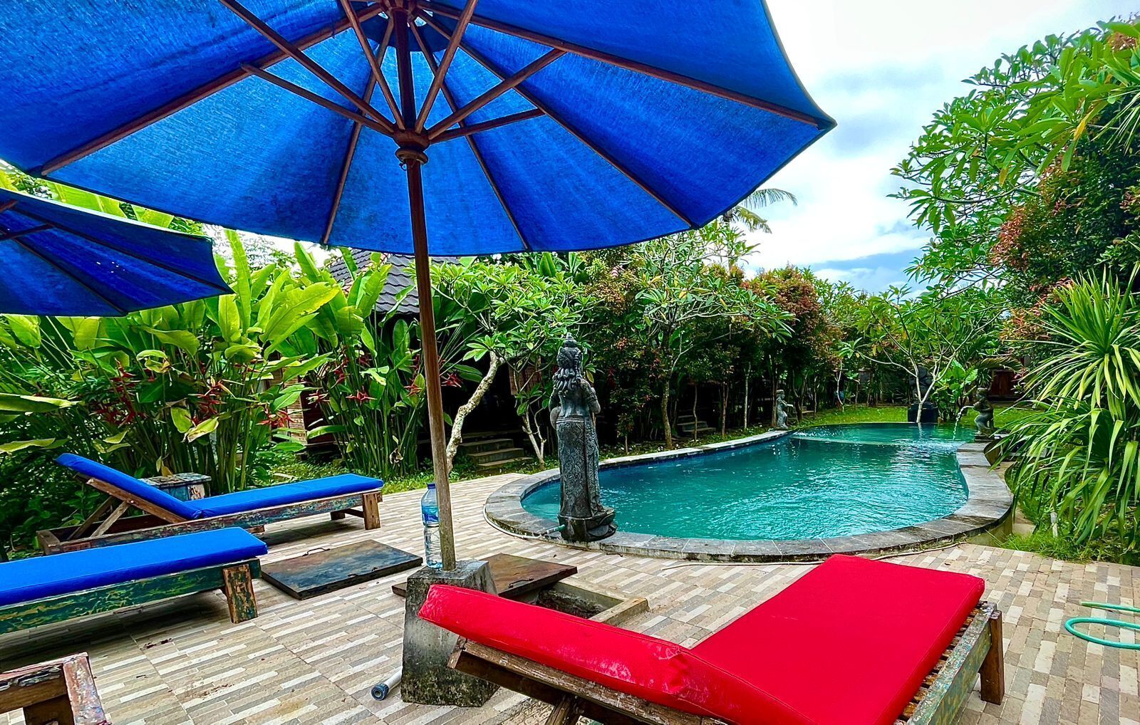 Zona de piscina con sillas de descanso azules y rojas, una sombrilla azul y una estatua.