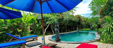 Zona de piscina con sillas de descanso azules y rojas, una sombrilla azul y una estatua.