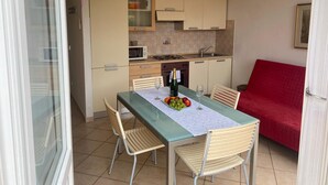 A compact kitchen with a dining area, including a table with chairs, a red sofa, and a glass door leading outside. - Green Oasis 1 (Lerici)