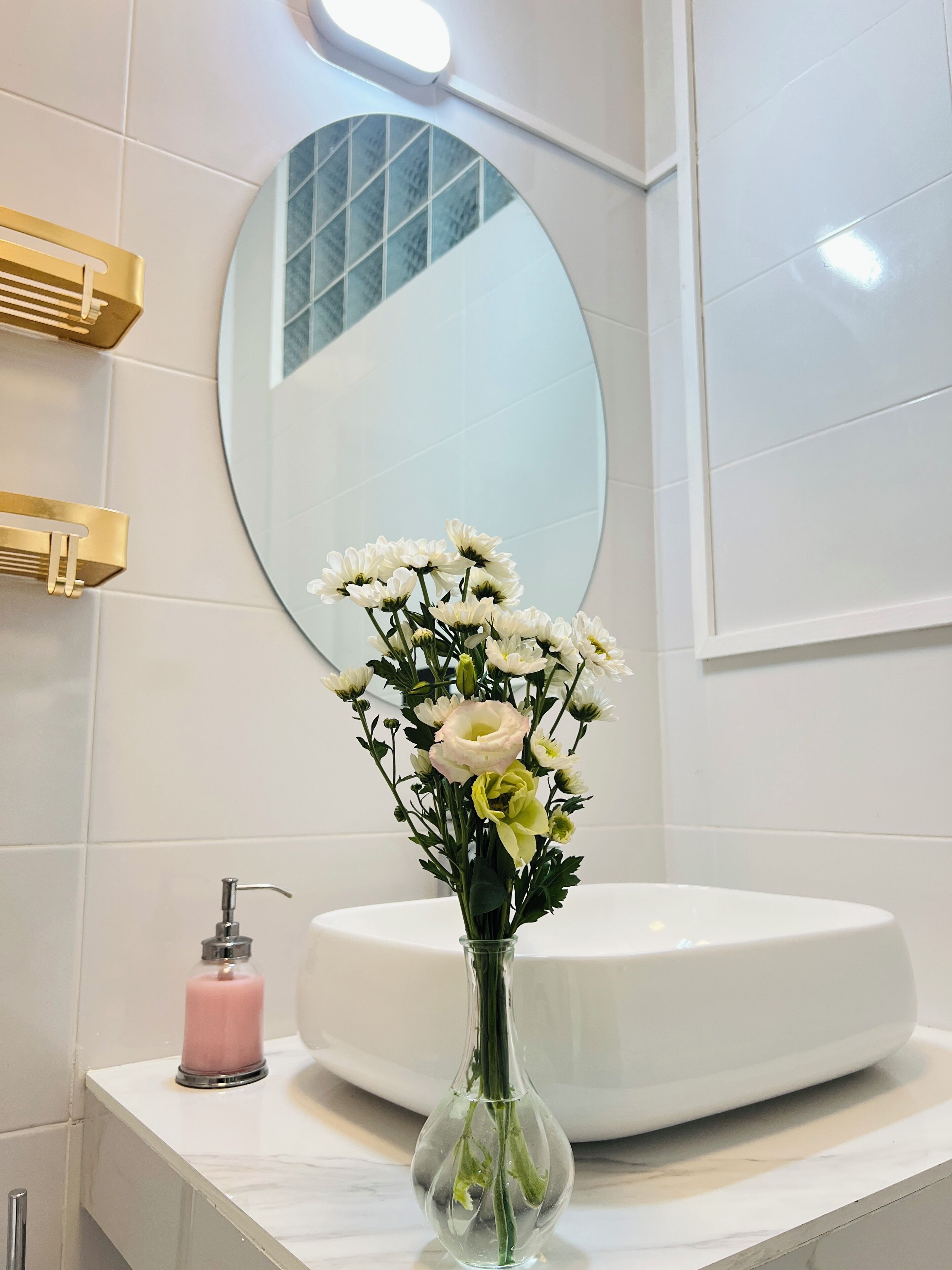 Um banheiro com pia branca, espelho redondo e um vaso com flores brancas.