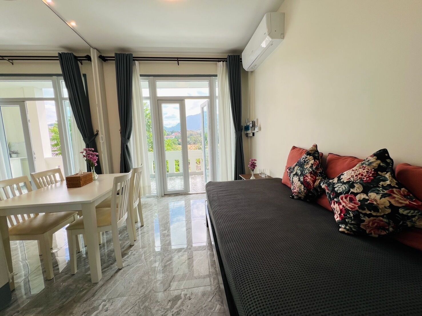 A hotel room with a bed, a dining table with chairs, and a balcony with curtains.