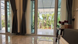 Ένα δωμάτιο ξενοδοχείου με μια μεγάλη συρόμενη γυάλινη πόρτα που οδηγεί σε έναν σκεπαστό εξωτερικό χώρο, ένα κρεβάτι με σκούρο προσκέφαλο και ένα μικρό τραπέζι με ένα βάζο γεμάτο κόκκινα τριαντάφυλλα.