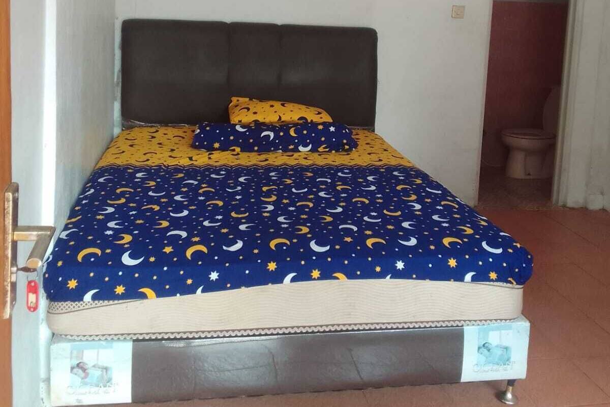 Una cama con colcha azul y amarilla estampada, cabecera oscura y un cuadro en la pared.