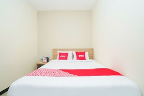 Una camera d'albergo con un letto, due cuscini con il logo "OYO", un comodino con una bottiglia e una scatola, e una coperta a motivi rossi e bianchi.