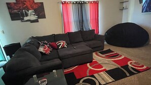 A living room with a sectional sofa, a glass coffee table, a red and black patterned rug, a ceiling fan, and a wall-mounted TV. - Charming 4-bedroom house with AC and WiFi in lovely Indianapolis (Indianapolis)