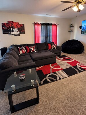 Un salon moderne avec un canapé d’angle, une table basse en verre, un fauteuil poire et un tapis aux motifs rouges et noirs.