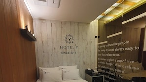 Ein Hotelzimmer mit einem Bett, einer an der Wand befestigten Ablagefläche mit Dekoration, einer Wand mit Logo und Text sowie einem Spiegel mit motivierenden Zitaten.