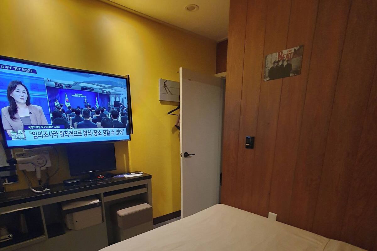 Habitación de hotel con cama, escritorio con televisor mostrando un noticiero, armario de madera y una puerta.