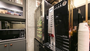 Eine Kaffeemaschine mit digitaler Anzeige und Tasten, ein Regal mit Büchern und ein Stapel weißer Tassen.