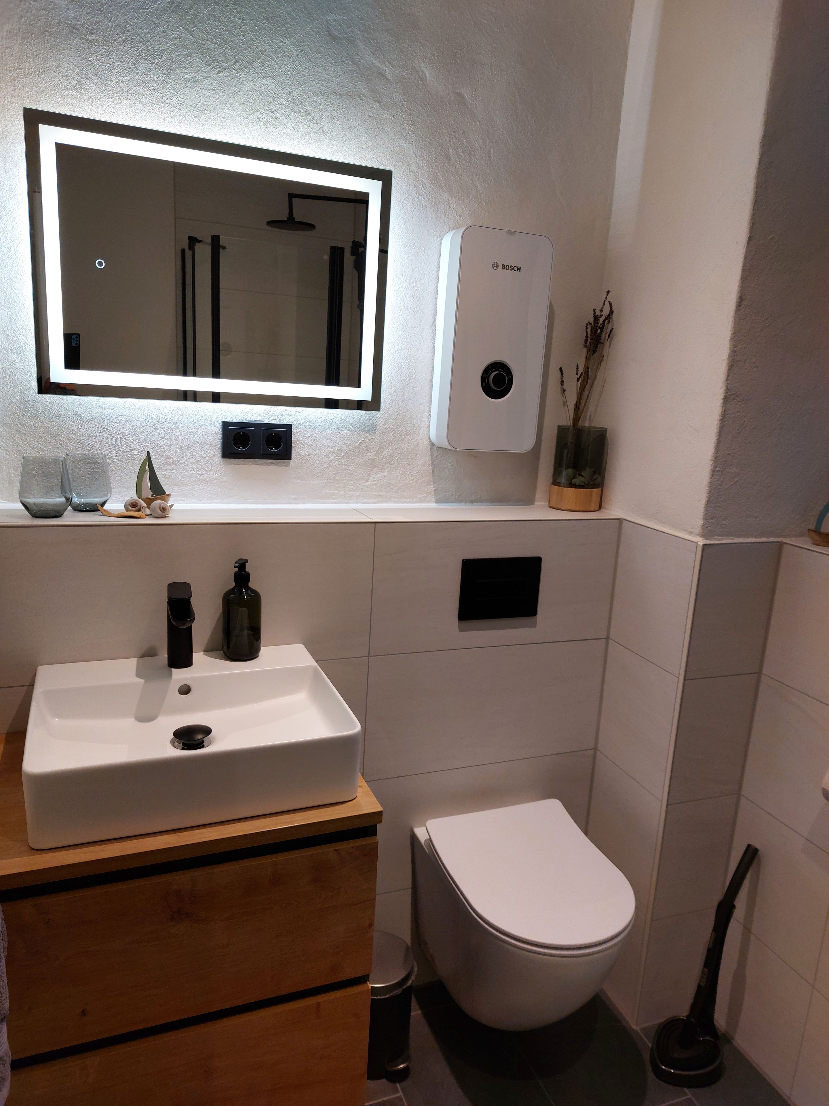 A bathroom with a white sink, a mirror, a Bosch water heater, a toilet, and a handheld vacuum cleaner.