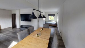 Un salon moderne avec une table à manger en bois, des canapés gris, un téléviseur à écran plat et un lit près d’une fenêtre.